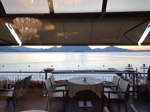 A serene lakeside view from a restaurant terrace, featuring elegantly set tables with white tablecloths and wicker chairs. The water reflects the soft light of a sunset, and distant mountains enhance the tranquil scenery. Chandeliers hang from the ceiling above, adding a touch of luxury.