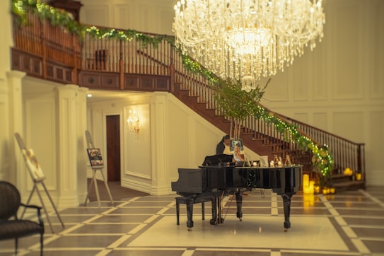 A grand staircase adorned with greenery and a large chandelier above it dominates the scene. Below, a black grand piano is positioned with music sheets and framed photos on it. The floor has a checkered pattern, and there are soft, warm lighting fixtures throughout the elegant setting.