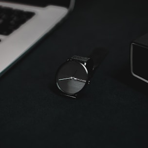 A sleek black luxury watch with minimalist design resting on a dark textured surface.