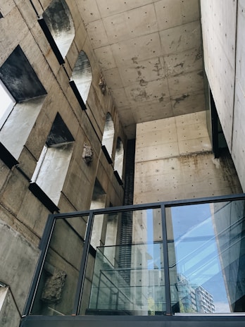 A modern architectural space featuring concrete walls with arched openings and a ceiling showing visible marks and stains. The design includes large glass windows and railings, creating a juxtaposition between the industrial materials and the sleek finish. The structure outside the window appears to be an urban skyline with a focus on geometric shapes.