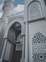 The image features a beautifully designed architectural structure with intricate geometric patterns on the window grills and archways. The building's exterior includes tall, ornate columns and a prominent, decorative minaret rising into a sky with scattered clouds.