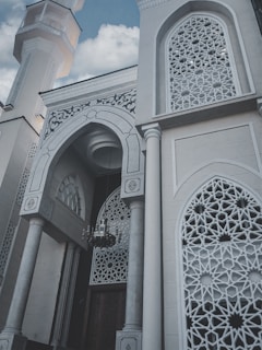 The image features a beautifully designed architectural structure with intricate geometric patterns on the window grills and archways. The building's exterior includes tall, ornate columns and a prominent, decorative minaret rising into a sky with scattered clouds.