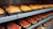 Slices of fruit, including cantaloupe and watermelon, are neatly arranged in plastic trays on metal shelves. The fruit is fresh and vibrant, with the cantaloupe appearing orange and the watermelon displaying deep red hues with green rinds. Nearby, cups filled with mixed fruit are visible.