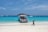 A speedboat is docked on a sandy beach with clear turquoise water. Several other boats are visible in the distance, and a person is walking along the shoreline. A small island covered with greenery is visible on the horizon under a clear blue sky.