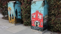 Painted utility boxes feature vibrant street art depicting two distinct scenes. One box displays an image of people, including a man and a woman, near a soldier under a blue sky. The other box shows a red building with windows and doors under a similarly stylized sky, with power lines running across.