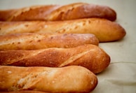 Several crusty baguettes are aligned on a light surface, showcasing their golden-brown, crispy exteriors. The loaves have a slightly shiny finish and display the characteristic elongated shape and slashes typical of French bread.