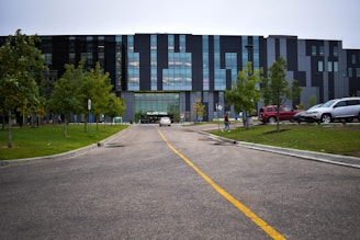 A modern building with a facade made of glass and dark panels is situated at the end of a road marked with a yellow dividing line. The area is surrounded by evenly spaced trees and neatly trimmed grass. A few vehicles are parked on the right side, and a person is walking towards the building entrance, which is labeled Allard Hall.