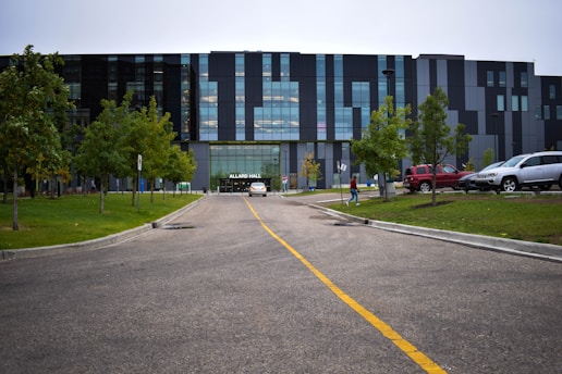A modern building with a facade made of glass and dark panels is situated at the end of a road marked with a yellow dividing line. The area is surrounded by evenly spaced trees and neatly trimmed grass. A few vehicles are parked on the right side, and a person is walking towards the building entrance, which is labeled Allard Hall.
