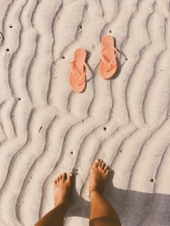 A pair of orange flip-flops placed on a sandy beach with wavy patterns. Bare feet with a small anklet can be seen at the bottom of the image, capturing a relaxed and casual vibe in a beach setting.