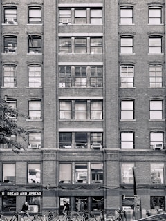 A multi-story brick building with numerous windows, some of which have air conditioning units installed. The ground floor has a storefront named 'BREAD AND SPREAD' with people walking past. Bicycles are parked in racks along the sidewalk.