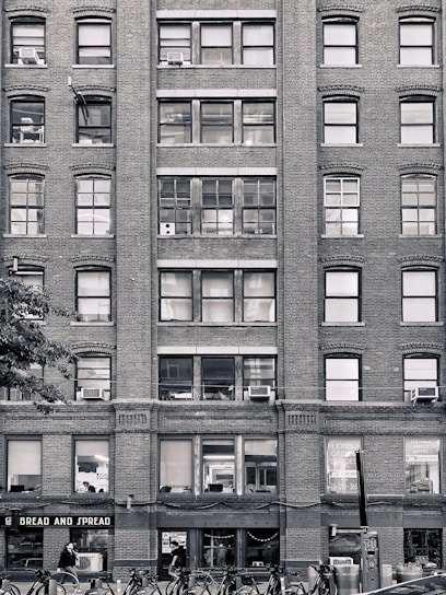A multi-story brick building with numerous windows, some of which have air conditioning units installed. The ground floor has a storefront named 'BREAD AND SPREAD' with people walking past. Bicycles are parked in racks along the sidewalk.