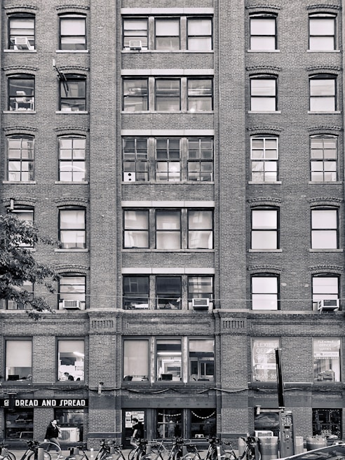 A multi-story brick building with numerous windows, some of which have air conditioning units installed. The ground floor has a storefront named 'BREAD AND SPREAD' with people walking past. Bicycles are parked in racks along the sidewalk.