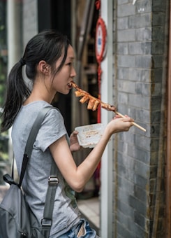 A young woman is eating from a skewer with chicken feet while holding an empty plastic container in her other hand. She stands outside a building with gray brick walls, wearing a gray T-shirt and a backpack.