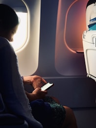 A person is seated next to an airplane window, looking at a smartphone. The natural light from the window illuminates the person's face and hand. The seat-back pocket in front of them contains safety information cards.
