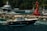 A luxurious motorboat with passengers is moving through clear blue water near a rocky shore and a red buoy. Several other boats and a large yacht are anchored near the docks in the background, surrounded by verdant trees.