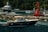 A luxurious motorboat with passengers is moving through clear blue water near a rocky shore and a red buoy. Several other boats and a large yacht are anchored near the docks in the background, surrounded by verdant trees.