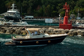 A luxurious motorboat with passengers is moving through clear blue water near a rocky shore and a red buoy. Several other boats and a large yacht are anchored near the docks in the background, surrounded by verdant trees.