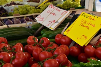 A vibrant display of fresh produce at a market stall, with red tomatoes in the foreground, green cucumbers, and bunches of grapes and figs in the background. There are signs with handwritten notes indicating prices and descriptions.
