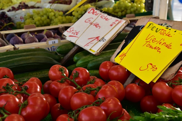 A vibrant display of fresh produce at a market stall, with red tomatoes in the foreground, green cucumbers, and bunches of grapes and figs in the background. There are signs with handwritten notes indicating prices and descriptions.