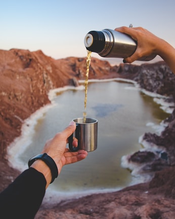 A person pours liquid from a thermos into a metal cup with a scenic backdrop of a lake surrounded by rocky terrain. The scene captures a sense of tranquility and outdoor adventure.