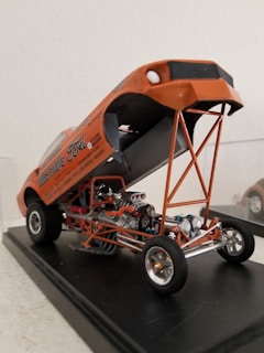 A detailed miniature model of a vintage drag racing car is displayed with its orange body lifted to reveal the intricate details of the engine and chassis. The car is positioned on a black platform, showcasing the metallic engine components, exposed piping, and rubber wheels.
