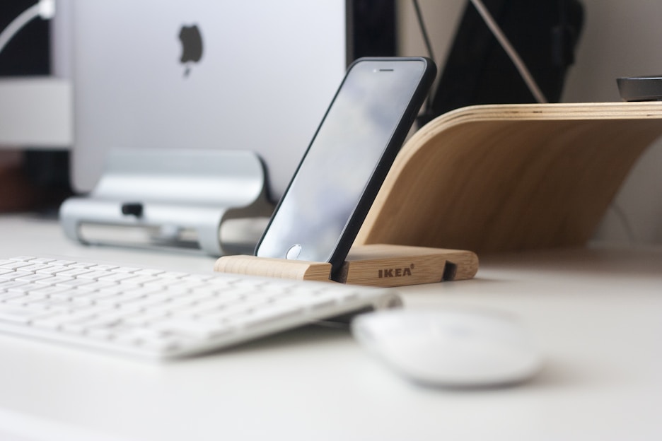 A sleek adjustable phone holder on a desk holding a smartphone at eye level with a tablet beside it.