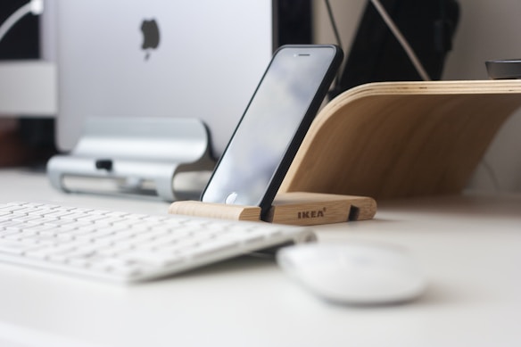 An organized workspace featuring a mounted smartphone on a wooden stand, positioned in front of a desktop computer. A wireless keyboard and mouse are placed nearby, all set on a clean, white desk. A wooden curve-shaped document holder is also visible, complementing the minimalist and modern aesthetic.