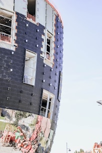 A modern building under construction with a curved facade is wrapped in black materials and features reflective lower panels. Openings for windows and evidence of construction materials are visible, with scaffolding and a crane appearing in the surroundings.