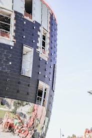 A modern building under construction with a curved facade is wrapped in black materials and features reflective lower panels. Openings for windows and evidence of construction materials are visible, with scaffolding and a crane appearing in the surroundings.