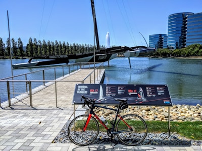 A catamaran is displayed prominently over a body of water, supported by structures, with its name and achievements visible. In the foreground, a bicycle is parked on a paved walkway beside signage detailing statistics. To the right, modern glass buildings rise, contrasting with the natural background of trees and a water fountain.