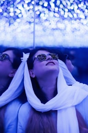 A person is looking upwards with a joyful expression, surrounded by shimmering, blue lights. They are wearing sunglasses that reflect the lights, and a white scarf drapes elegantly over their shoulders. The background consists of numerous small, blurred lights that create a dreamy, ethereal atmosphere.
