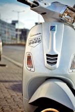 Close-up view of a white scooter parked on the side of a road. The scooter has a number plate with the registration MH 12 QX 6622. The background shows a blurred urban environment with the hint of parked cars and a building.