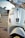Close-up view of a white scooter parked on the side of a road. The scooter has a number plate with the registration MH 12 QX 6622. The background shows a blurred urban environment with the hint of parked cars and a building.