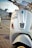 Close-up view of a white scooter parked on the side of a road. The scooter has a number plate with the registration MH 12 QX 6622. The background shows a blurred urban environment with the hint of parked cars and a building.
