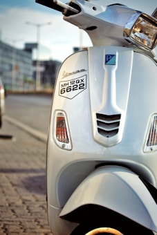 Close-up view of a white scooter parked on the side of a road. The scooter has a number plate with the registration MH 12 QX 6622. The background shows a blurred urban environment with the hint of parked cars and a building.
