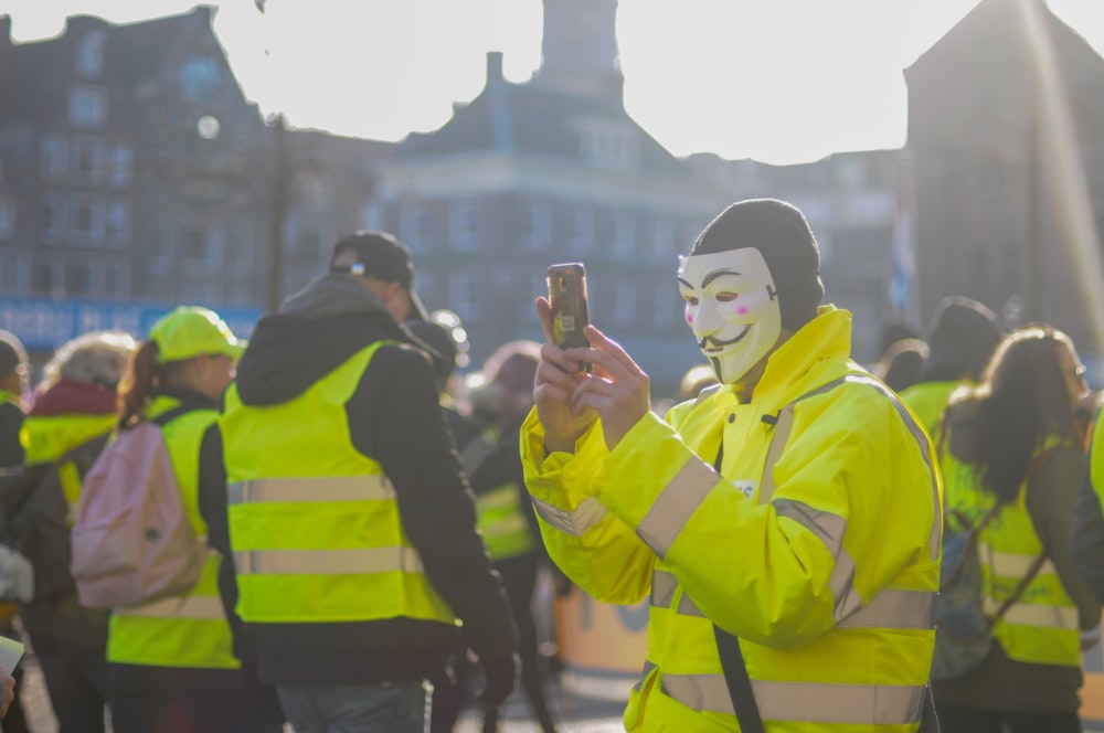 Person mit Guy-Fawkes-Maske mit Telefon umgeben von Menschen