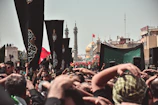 A large crowd gathers for a religious event, with many people raising their hands in a display of devotion. Black flags with ornate designs are prominently displayed among the crowd. In the background, the golden dome of a significant mosque or shrine and tall minarets can be seen, along with other structures and a clear sky.
