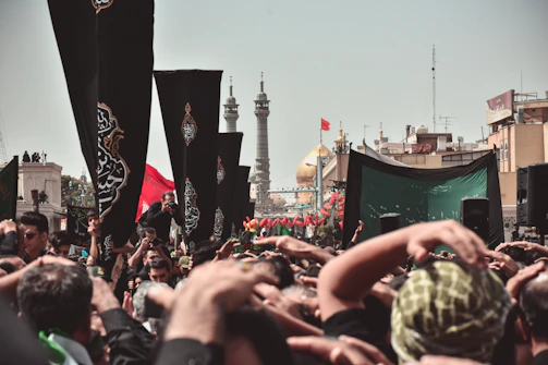 A large crowd gathers for a religious event, with many people raising their hands in a display of devotion. Black flags with ornate designs are prominently displayed among the crowd. In the background, the golden dome of a significant mosque or shrine and tall minarets can be seen, along with other structures and a clear sky.