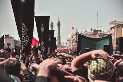 A large crowd gathers for a religious event, with many people raising their hands in a display of devotion. Black flags with ornate designs are prominently displayed among the crowd. In the background, the golden dome of a significant mosque or shrine and tall minarets can be seen, along with other structures and a clear sky.