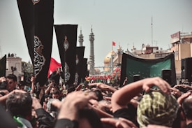 A large crowd gathers for a religious event, with many people raising their hands in a display of devotion. Black flags with ornate designs are prominently displayed among the crowd. In the background, the golden dome of a significant mosque or shrine and tall minarets can be seen, along with other structures and a clear sky.