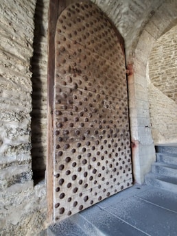 A large, heavy wooden door reinforced with numerous metal studs is inset into a stone wall. The texture of the stones and the pattern of the studs on the door create a historical and medieval appearance. The door is partially open, leading to a stone walkway.