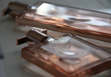 Two rectangular glass perfume bottles are lying on a reflective surface. The bottles have a sleek design with metallic caps and appear to be filled with a light peach-colored liquid, which reflects light softly.