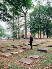 A person stands in a cemetery surrounded by gravestones, with tall trees overhead. The ground is covered with fallen autumn leaves, and bright brick buildings are visible in the background beyond an iron fence.