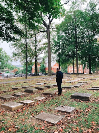 A person stands in a cemetery surrounded by gravestones, with tall trees overhead. The ground is covered with fallen autumn leaves, and bright brick buildings are visible in the background beyond an iron fence.