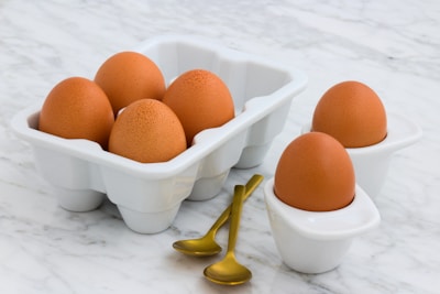 Six brown eggs are neatly placed in white ceramic trays on a marble surface, accompanied by two small, gold-colored spoons.