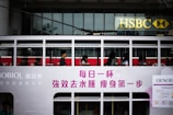 Passengers are seated inside a white, double-decker tram with advertisements and writing in Chinese characters on the side. The tram is passing by an HSBC bank building with its recognizable logo visible in the background.