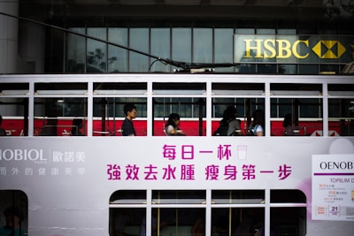 Passengers are seated inside a white, double-decker tram with advertisements and writing in Chinese characters on the side. The tram is passing by an HSBC bank building with its recognizable logo visible in the background.