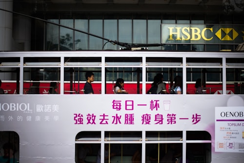 Passengers are seated inside a white, double-decker tram with advertisements and writing in Chinese characters on the side. The tram is passing by an HSBC bank building with its recognizable logo visible in the background.