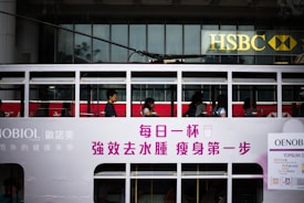 Passengers are seated inside a white, double-decker tram with advertisements and writing in Chinese characters on the side. The tram is passing by an HSBC bank building with its recognizable logo visible in the background.