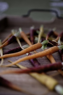 A collection of small, multicolored carrots scattered across a wooden surface. The focus is on the vibrant orange and purple hues of the carrots, with some green tops still attached. The wooden surface adds a rustic and earthy feel to the setting.